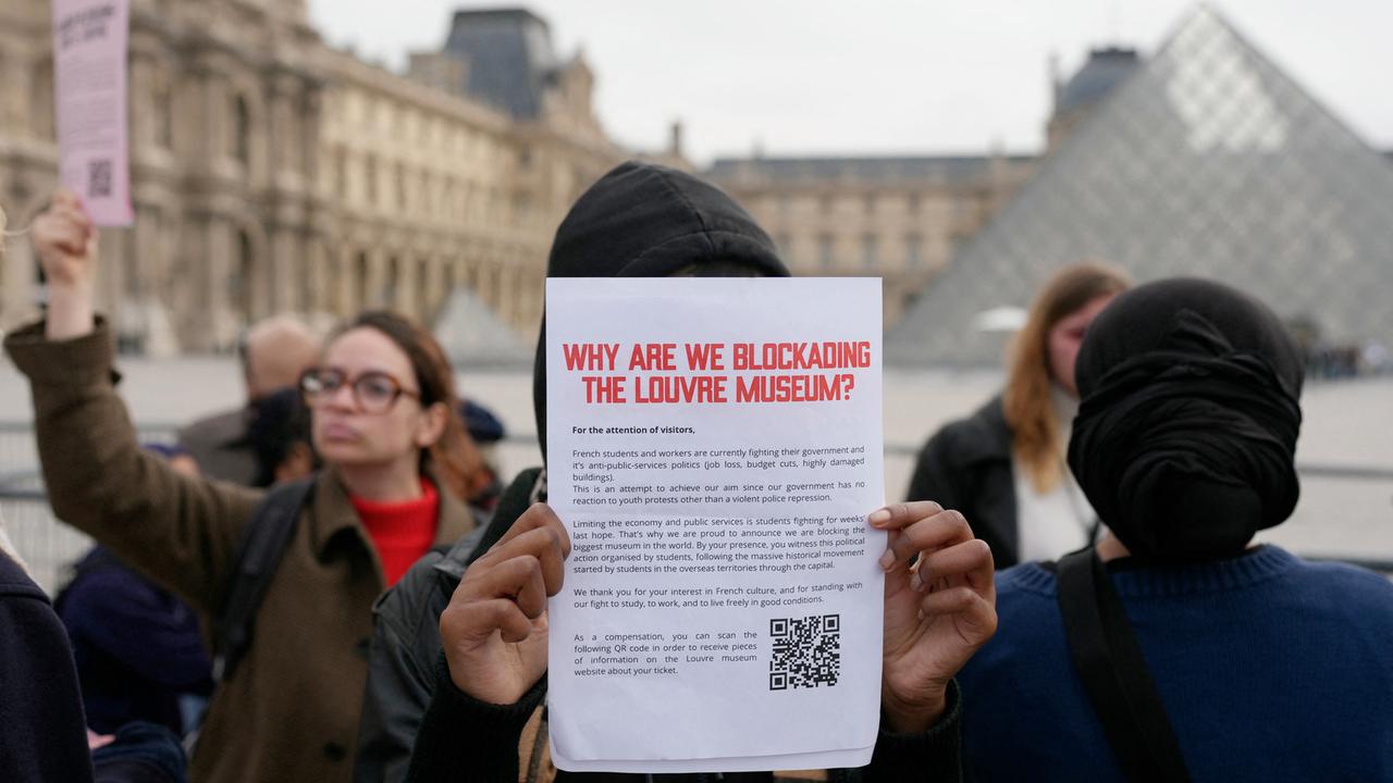 Eine demonstrierende Person hält vor dem Pariser Museum einen Zettel mit der Überschrift "Why are we blockading the Louvre Museum?" (auf Deutsch: "Warum blockieren wir den Louvre?") in die Kamera.
