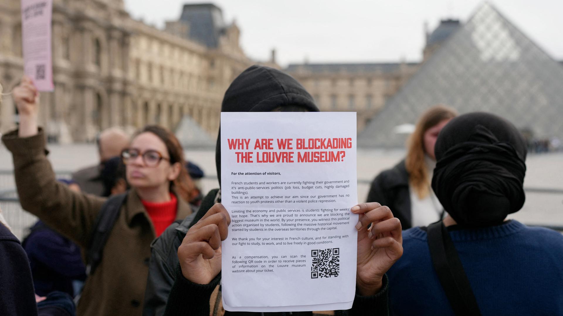 Eine demonstrierende Person hält vor dem Pariser Museum einen Zettel mit der Überschrift "Why are we blockading the Louvre Museum?" (auf Deutsch: "Warum blockieren wir den Louvre?") in die Kamera.