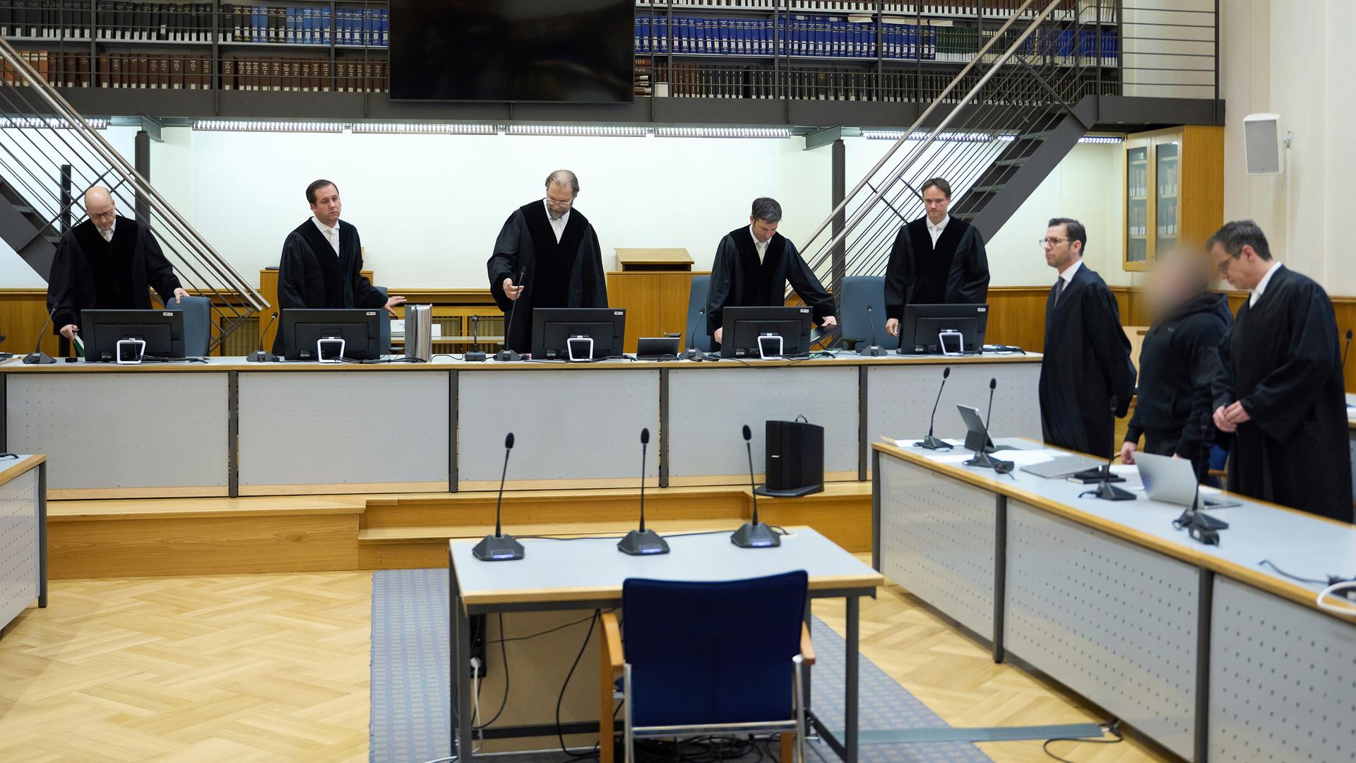 Blick in den Gerichtsaal: Hinter einem langen Tisch und einem kürzeren an der Seite stehen Männer in schwarzen Roben. 