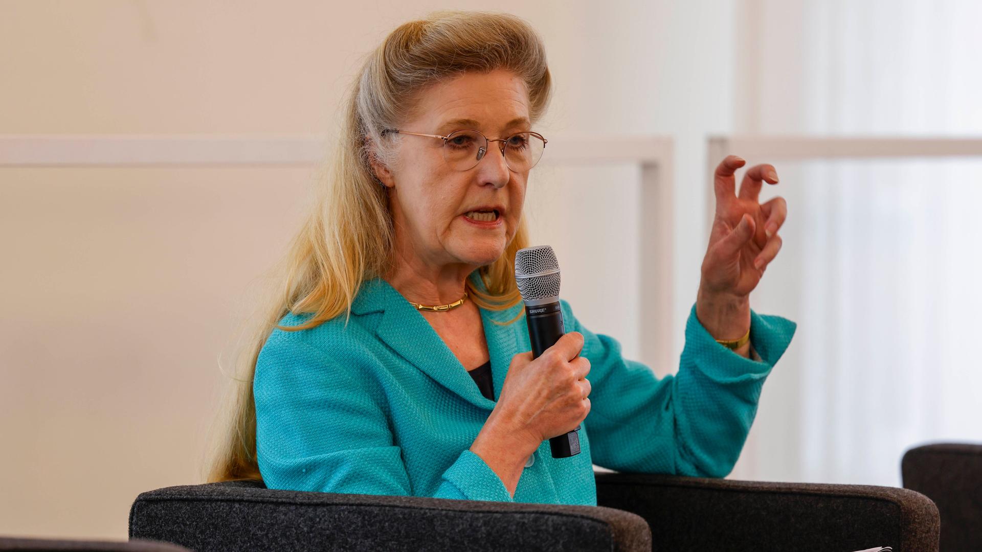 Eine bonde Frau mit Brille sitzt in einem Sessel. Sie hält sich ein Mikrofon vor den Mund und gestikuliert mit der anderen Hand. 