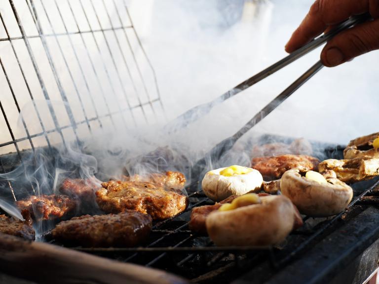 Aus einem dampfenden Grill liegen Gemüse- und Fleischstücke, das gerade mit einer Zange gewendet wird.
