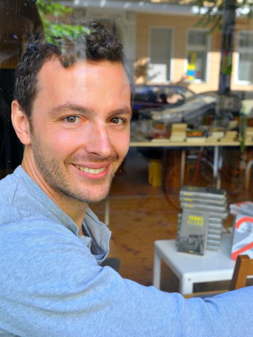 Der Verleger Sebastian Guggolz sitzt in der Geschäftsstelle des Guggolz-Verlags in Berlin-Schöneberg, im Hintergrund Bücherstapel. 