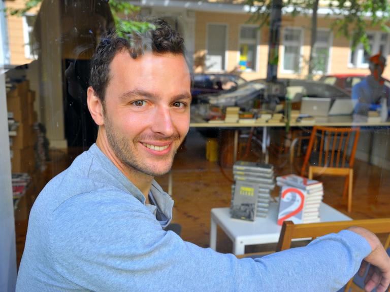 Der Verleger Sebastian Guggolz sitzt in der Geschäftsstelle des Guggolz-Verlags in Berlin-Schöneberg, im Hintergrund Bücherstapel. 