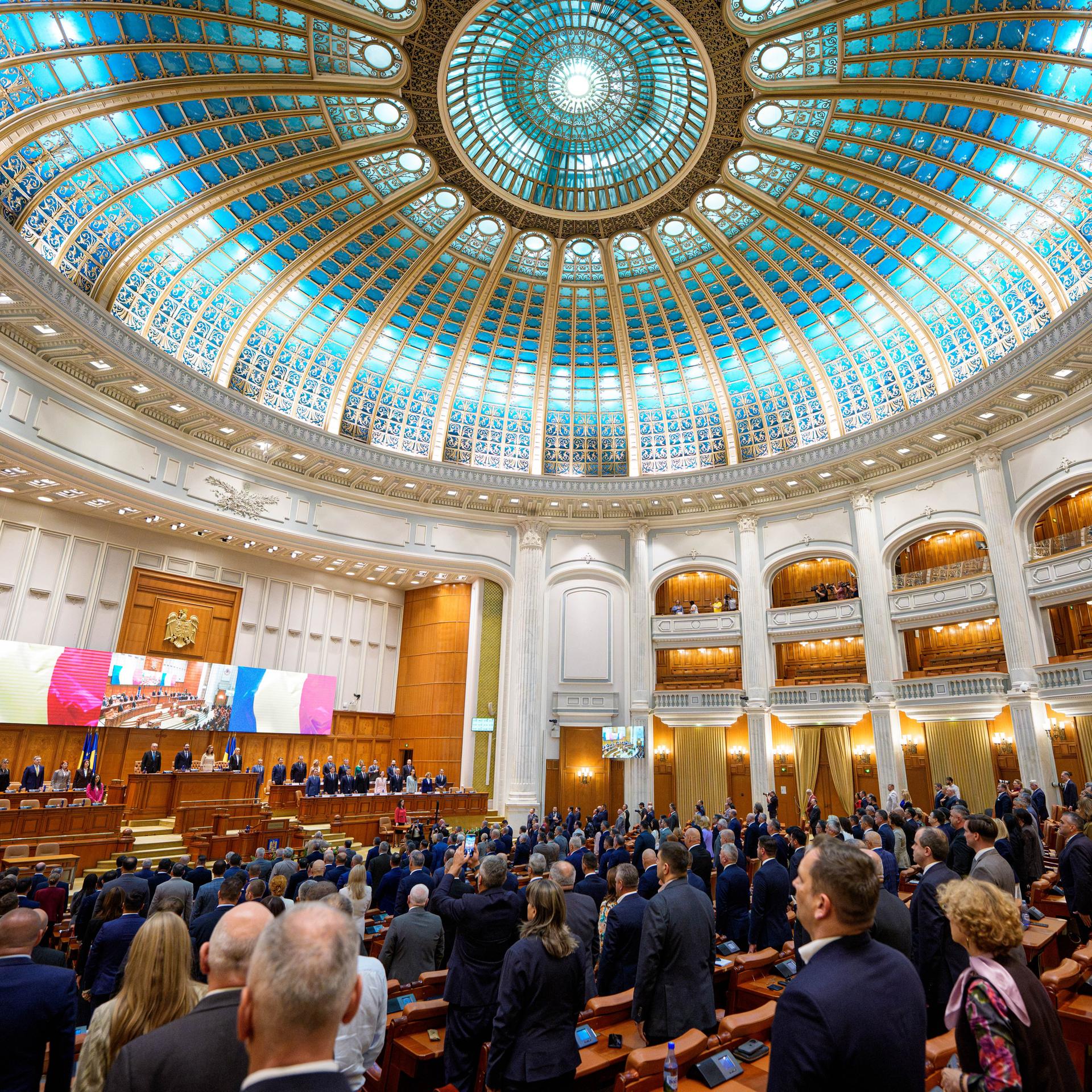 Blick in das vollbesetzte Parlament mit einer blauen Glaskuppel im Art-Deco-Stil. Die Abgeordneten stehen. 