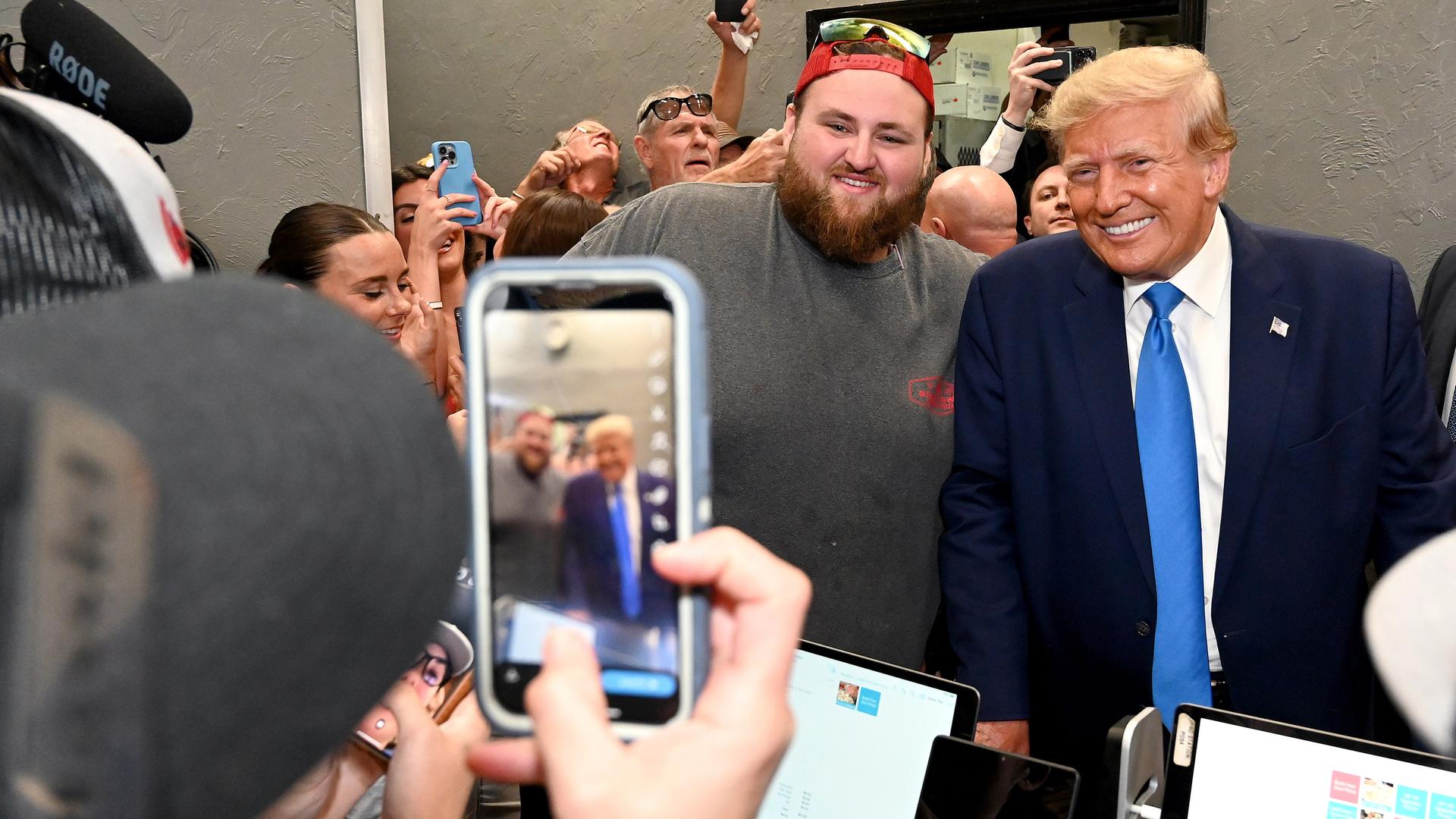 Ex-Präsident Trump und ein bärtiger Anhänger, der eine umgedrehte Baseballkappe und eine Sonnenbrille auf dem Kopf hat, lächeln in eine Smartphone Kamera.