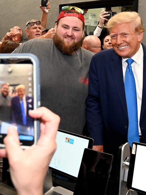 Ex-Präsident Trump und ein bärtiger Anhänger, der eine umgedrehte Baseballkappe und eine Sonnenbrille auf dem Kopf hat, lächeln in eine Smartphone Kamera.