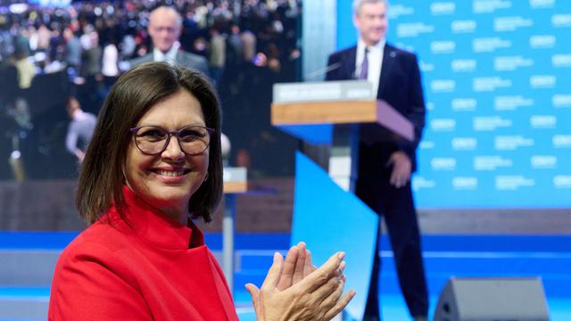 llse Aigner klatscht und blickt in die Kamera. Hinter ihr auf einer Bühne stehen Bundeskanzler Friedrich Merz und CSU-Chef Markus Söder.