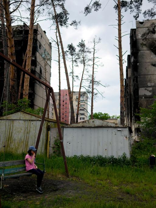 Ein Kind sitzt auf einer Schaukel in der ukrainischen Stadt Irpin. Im Hintergrund sind durch den Krieg zerstörte Wohngebäude zu sehen.
