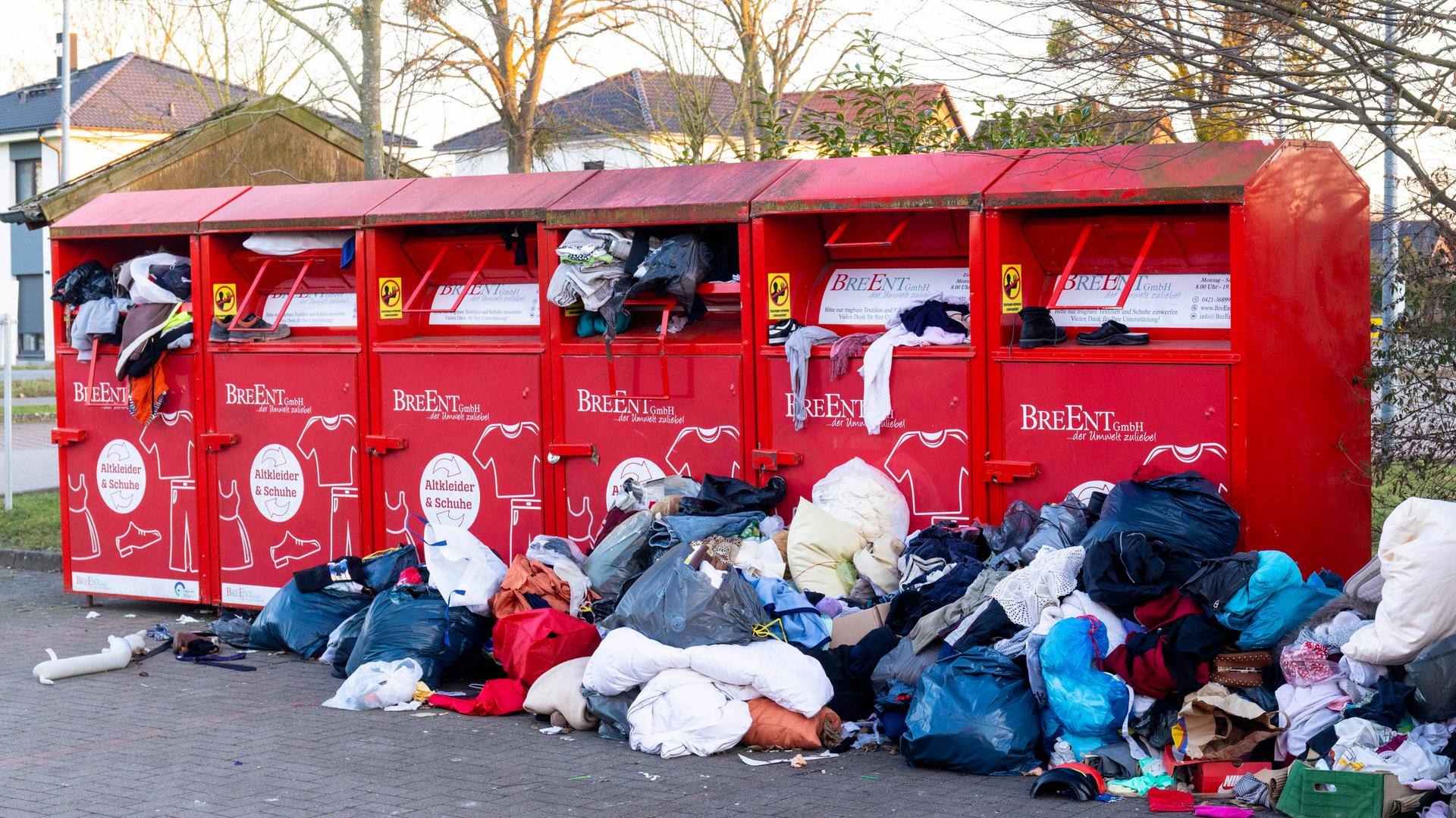 Rote Altkleidercontainer stehen auf einem Parkplatz. Sie quellen mit Kleidung über. Davor liegen Haufen aus Kleidung, die nicht mehr in die Container passen.