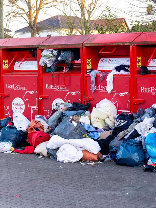 Rote Altkleidercontainer stehen auf einem Parkplatz. Sie quellen mit Kleidung über. Davor liegen Haufen aus Kleidung, die nicht mehr in die Container passen.