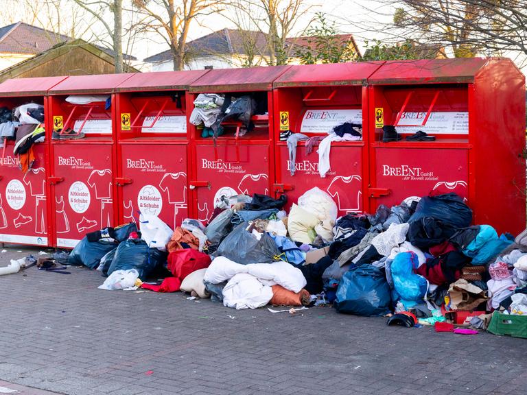 Rote Altkleidercontainer stehen auf einem Parkplatz. Sie quellen mit Kleidung über. Davor liegen Haufen aus Kleidung, die nicht mehr in die Container passen.