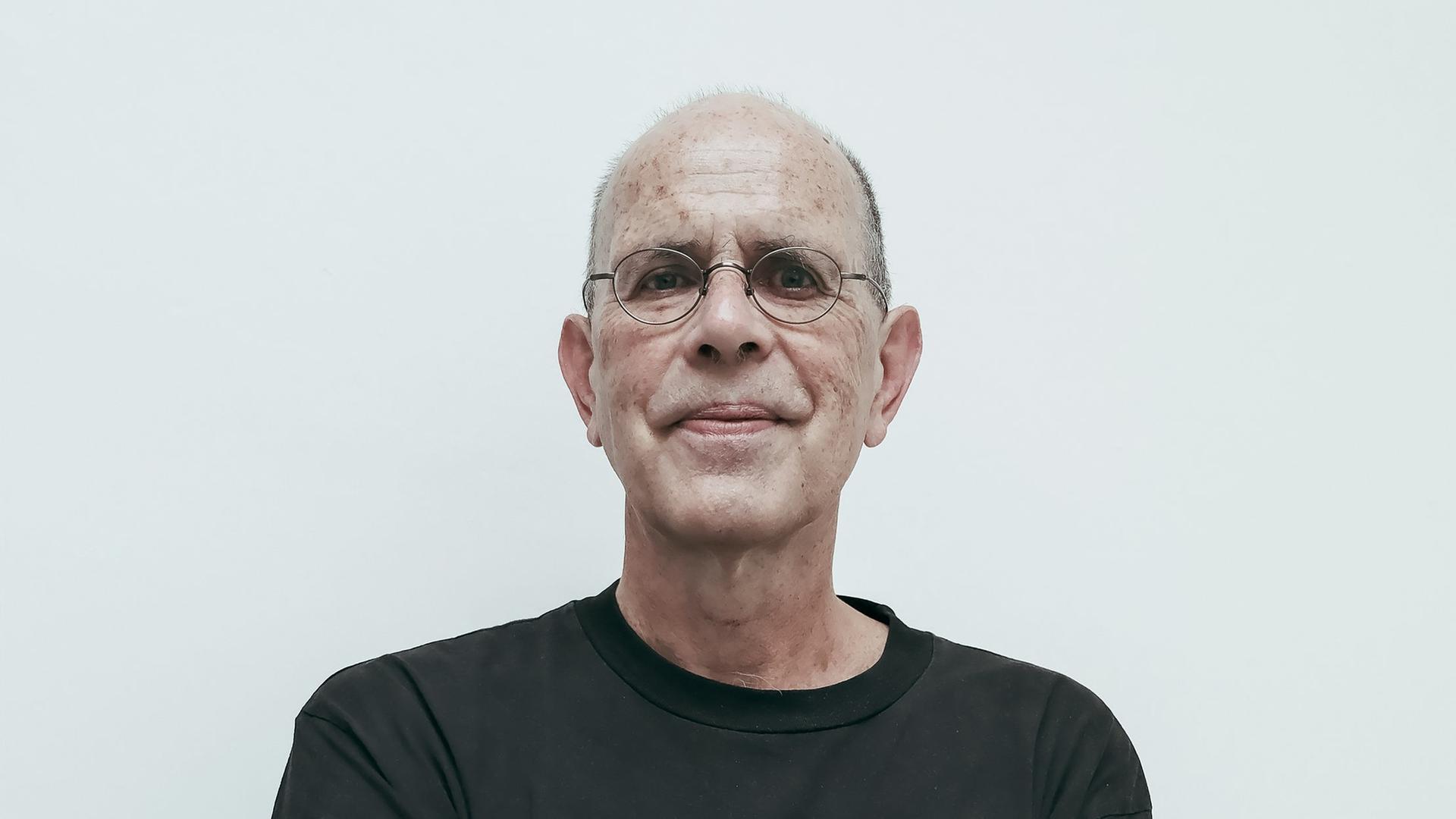 Ein Mann mit schwarzem Shirt und Brille verschränkt die Hände unter den Achseln udn schaut direkt in die Kamera