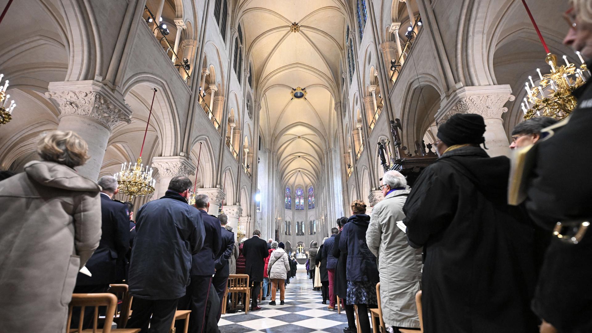 Blick in einen Gang von der Kathedrahle Notre Dame in Paris: Man sieht das neue Dach. Und unten sind Menschen in dicken Jacken. Sie blicken nach vorne zum Altar. Sie sind von ihren Stühlen aufgestanden. Blick in einen Gang von der Kathedrahle Notre Dame in Paris: Man sieht das neue Dach. Und unten sind Menschen in dicken Jacken. Sie blicken nach vorne zum Altar. Sie sind von ihren Stühlen aufgestanden.