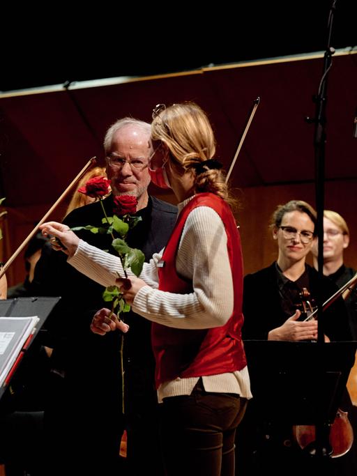 Eine rückwärtig zu sehende Frau mit Zopf verteilt Rosen an Orchester-Mitglieder, die ihre Instrumente in der Hand halten. Vor ihr stehen die Komponistin Raminta Serksnytë  und der Geiger Gidon Kremer.