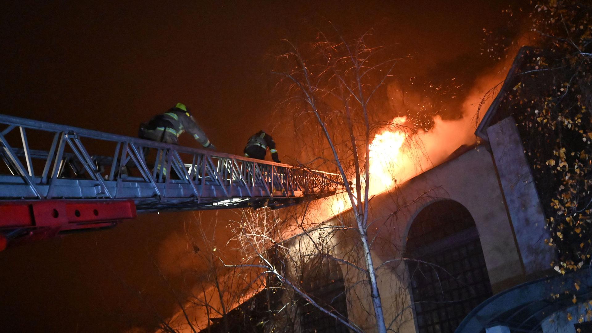 Feuerwehrmänner, stehen auf einer hohen Leiter und versuchen ein Feuer auf einem Haus zu löschen. Es ist Nacht.