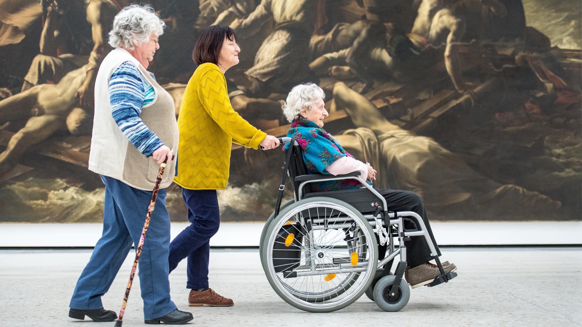 Drei ältere Damen gehen durch einen Ausstellungs-Raum im Städel-Museum. Eine sitzt im Rollstuhl und wird geschoben. Eine andere hat einen Gehstock.