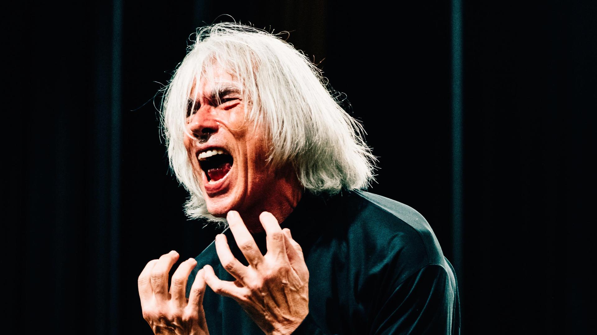 Ein Mann mit Schulterlangen weißen Haaren steht auf der Bühne und schreit, mit beiden Händen nach Ausdruck ringend
