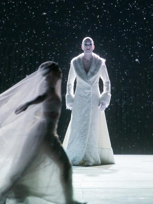 Bühnenfoto von der Aufführung "The Snow Queen". Darin steht ein Mann in einem pelzbesetzten weißen Mantel auf der Bühne. Um ihn herum tanzen verschleierte Frauen.