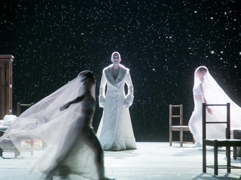Bühnenfoto von der Aufführung "The Snow Queen". Darin steht ein Mann in einem pelzbesetzten weißen Mantel auf der Bühne. Um ihn herum tanzen verschleierte Frauen.