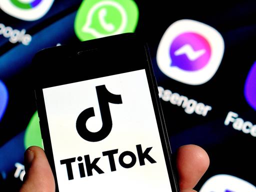 Eine Hand hält ein Smartphone mit einem Logo von TikTok vor dem Hintergrund eines weiteren Monitors mit Logos anderer Apps.