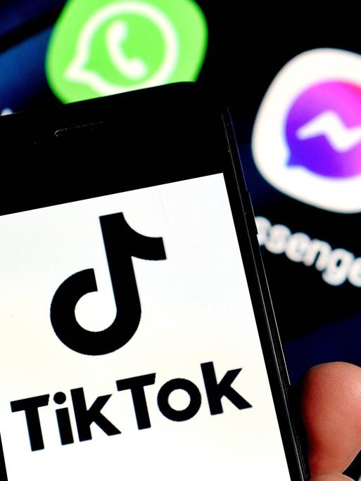 Eine Hand hält ein Smartphone mit einem Logo von TikTok vor dem Hintergrund eines weiteren Monitors mit Logos anderer Apps.