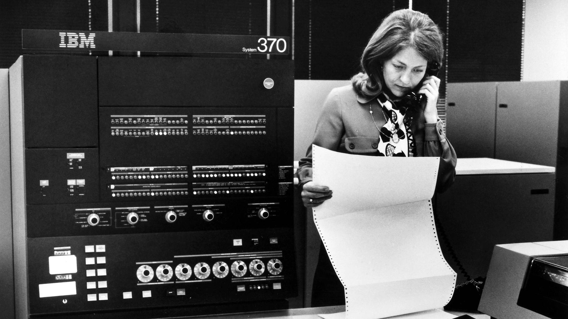 Auf einem Schwarz-Weiß-Foto ist der IBM-Großrechner System 370 zu sehen. Eine telefonierende Frau betrachtet einen Ausdruck. Auf einem Schwarz-Weiß-Foto ist der IBM-Großrechner System 370 zu sehen. Eine telefonierende Frau betrachtet einen Ausdruck.
