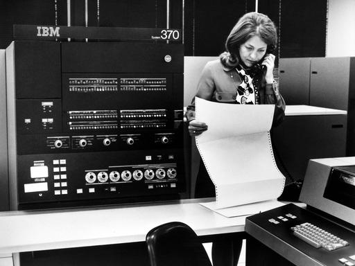 Auf einem Schwarz-Weiß-Foto ist der IBM-Großrechner System 370 zu sehen. Eine telefonierende Frau betrachtet einen Ausdruck.