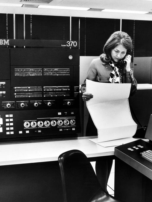 Auf einem Schwarz-Weiß-Foto ist der IBM-Großrechner System 370 zu sehen. Eine telefonierende Frau betrachtet einen Ausdruck.
