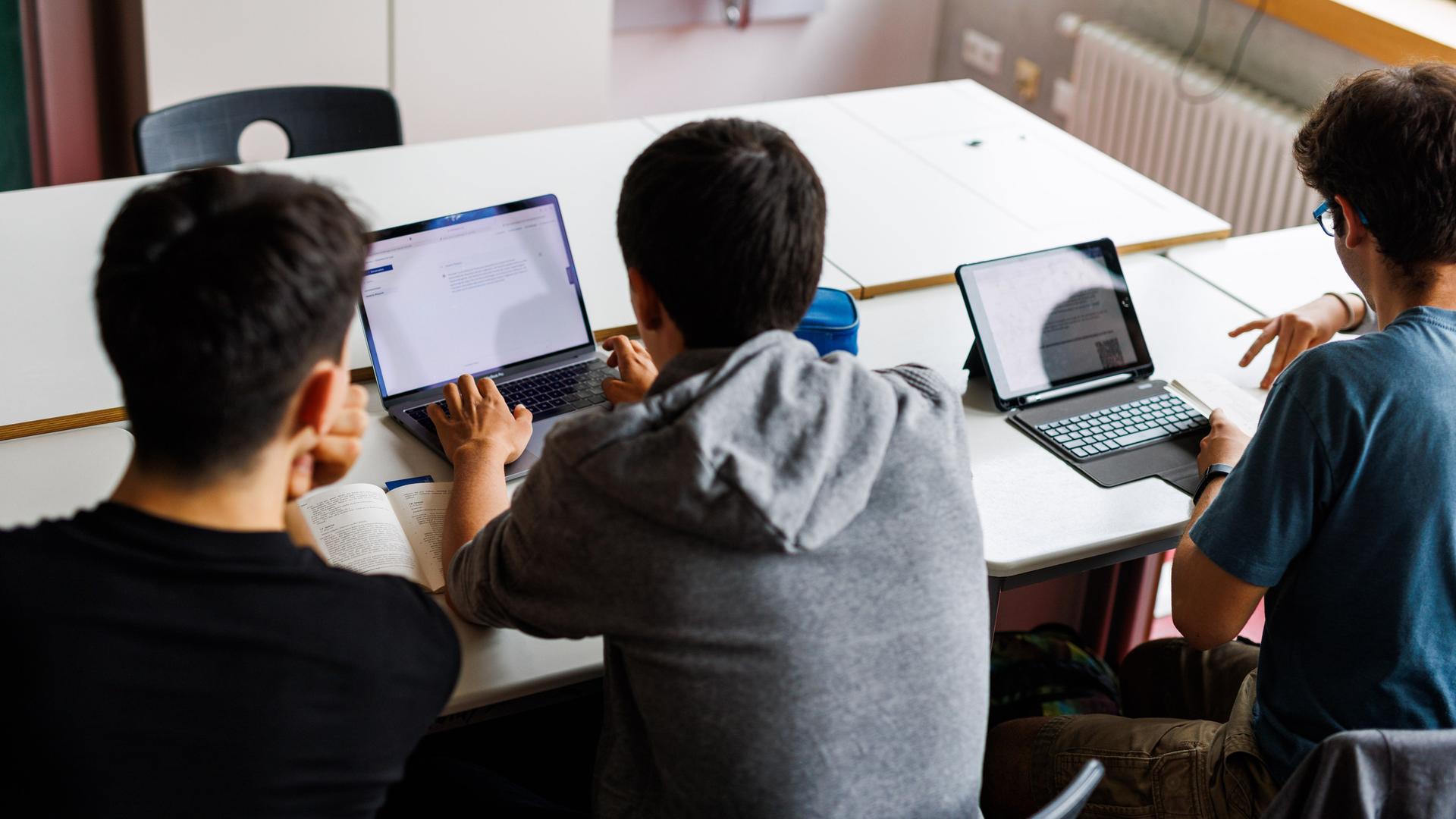 Schüler eines Gymnasiums sitzen vor einem Laptop und benutzen ein KI-Tool. Sie sind von hinten zu sehen und blicken auf ihre Laptops. 