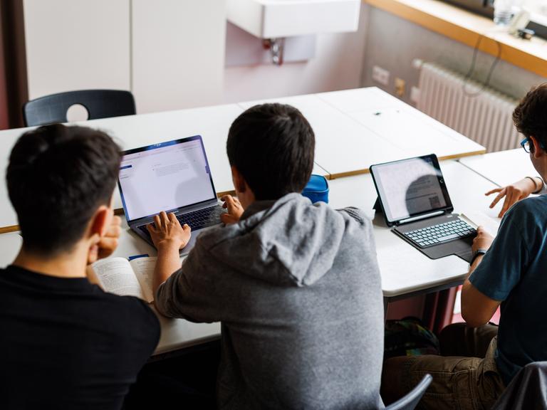 Schüler eines Gymnasiums sitzen vor einem Laptop und benutzen ein KI-Tool. Sie sind von hinten zu sehen und blicken auf ihre Laptops. 