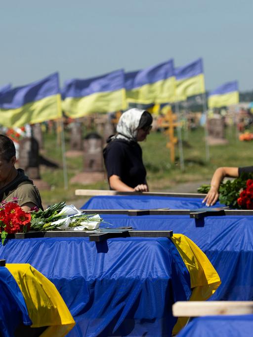 Angehörige und Freunde nehmen an der Begräbnisfeier für 13 an der Front gefallenen ukrainische Soldaten teil. Die Särge sind in einer Reihe aufgestellt und jeweils mit einer ukrainischen Fahne abgedeckt.