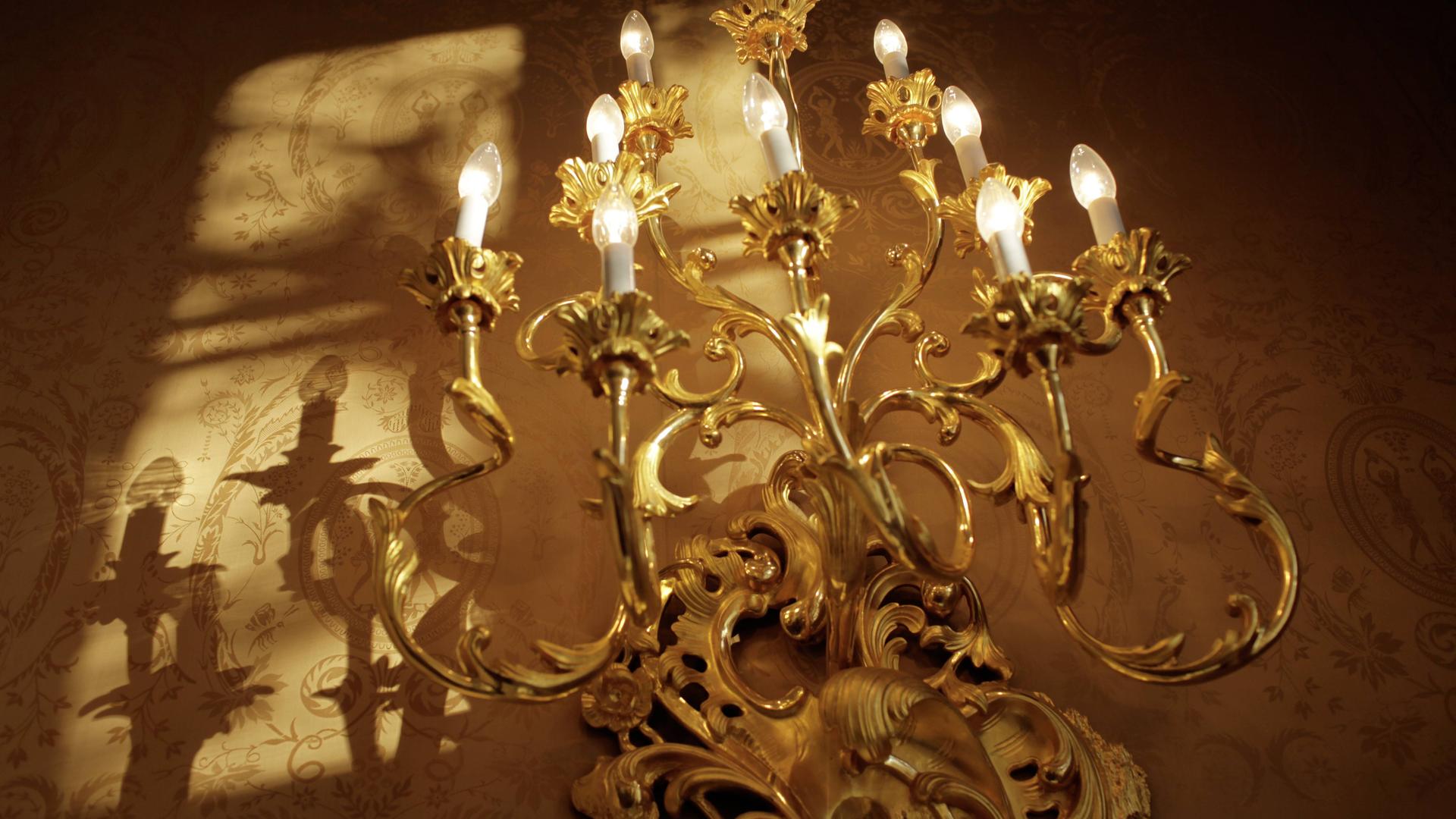 Ein goldener, barocker Wandleuchter hängt an der Wand und wirft neben ihm verschnörkelte Schatten.
