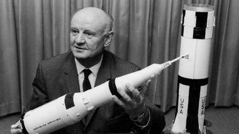 Ein kahlköpfiger Mann in Anzug und Krawatte sitzt an einem Tisch und hält das Modell einer Weltraumraktete in den Händen.