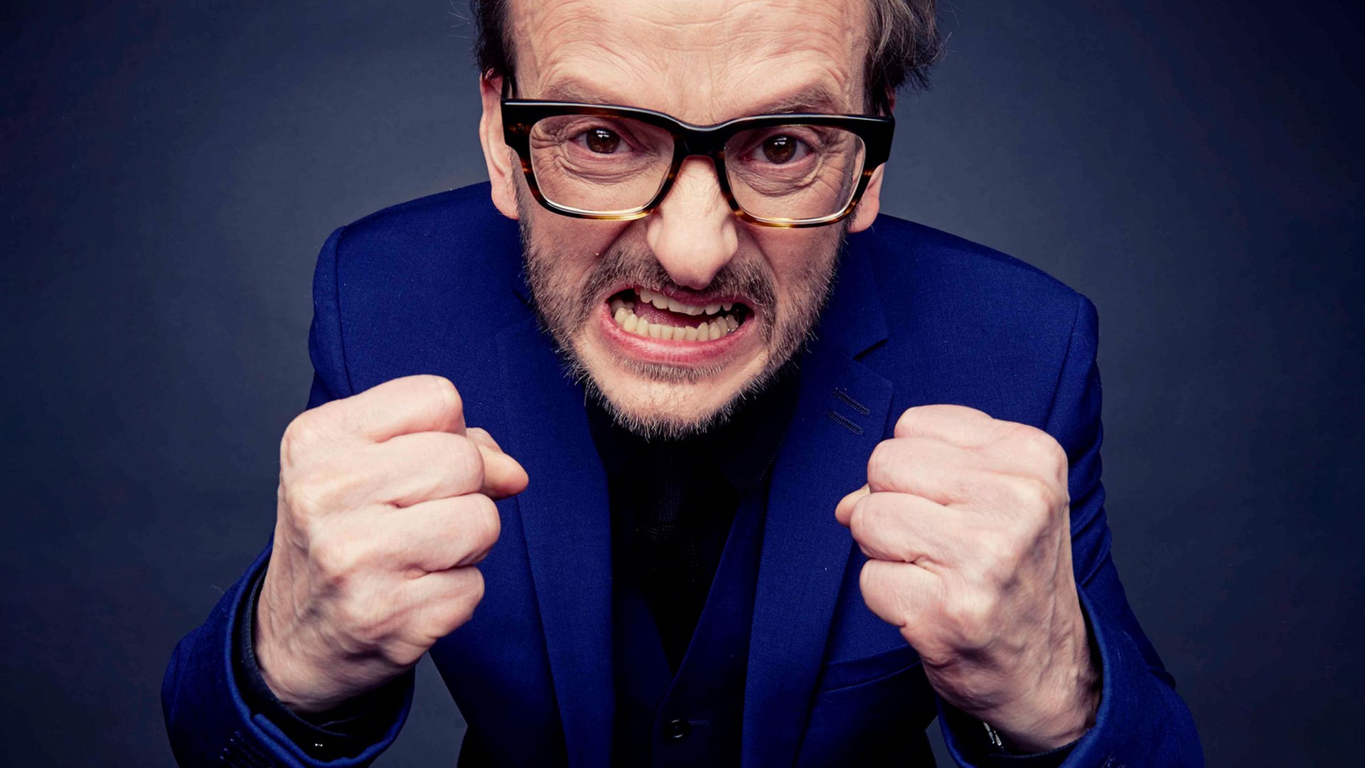 Portrait von Milan Peschel, der mit geballten Fäusten überzogen wütend in die Kamera schaut. Der Hintergrund ist dunkelblau. Portrait von Milan Peschel, der mit geballten Fäusten überzogen wütend in die Kamera schaut. Der Hintergrund ist dunkelblau.