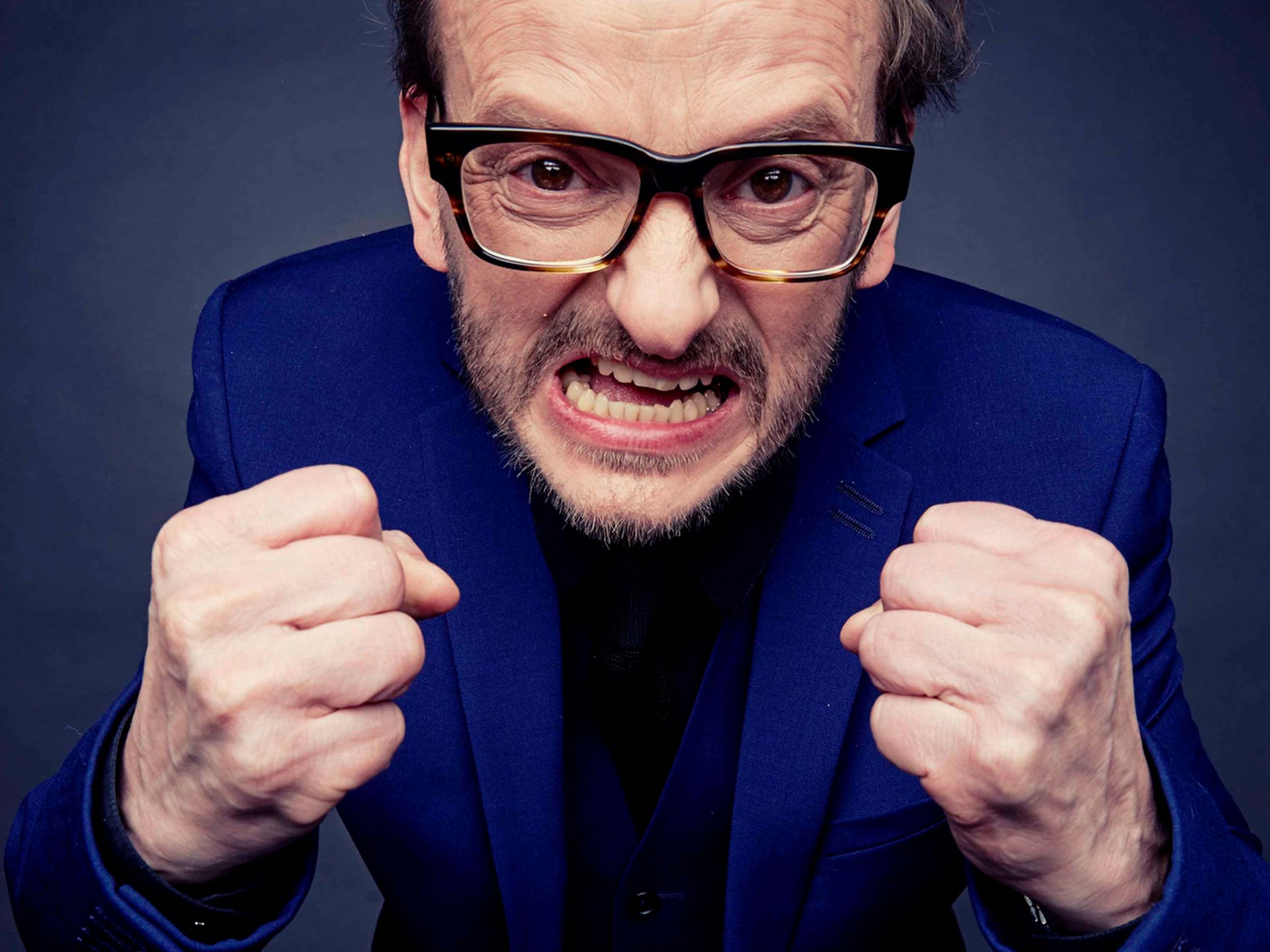Portrait von Milan Peschel, der mit geballten Fäusten überzogen wütend in die Kamera schaut. Der Hintergrund ist dunkelblau.