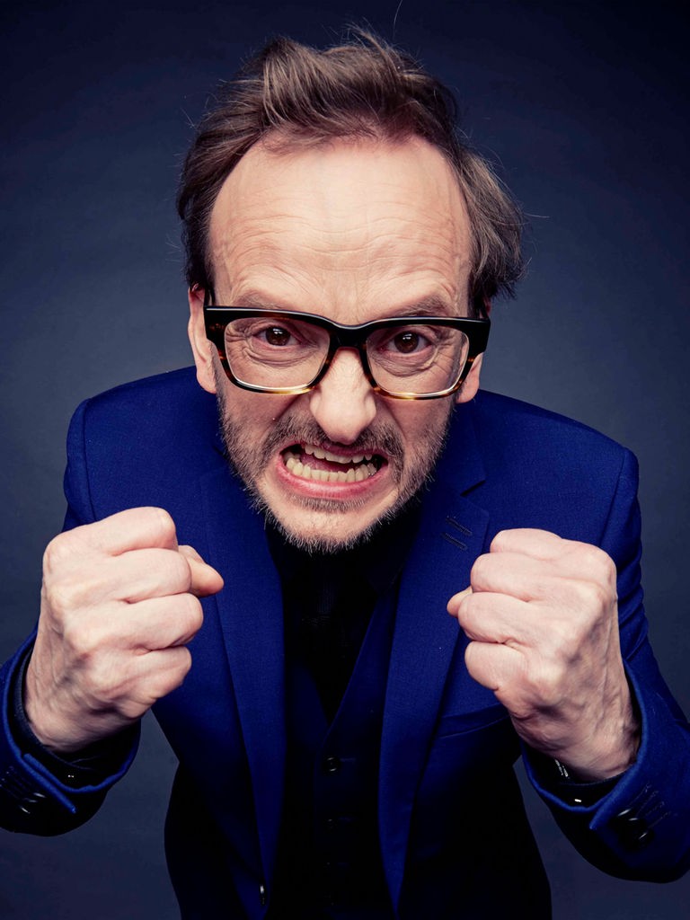 Portrait von Milan Peschel, der mit geballten Fäusten überzogen wütend in die Kamera schaut. Der Hintergrund ist dunkelblau.