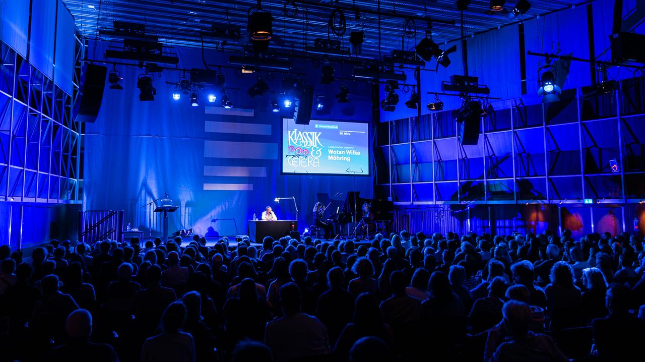 Ein Mann moderiert in einem blau beleuchteten Saal vor Publikum. Rechts von ihm ist ein Klaviertrio auf der Bühne zu sehen.