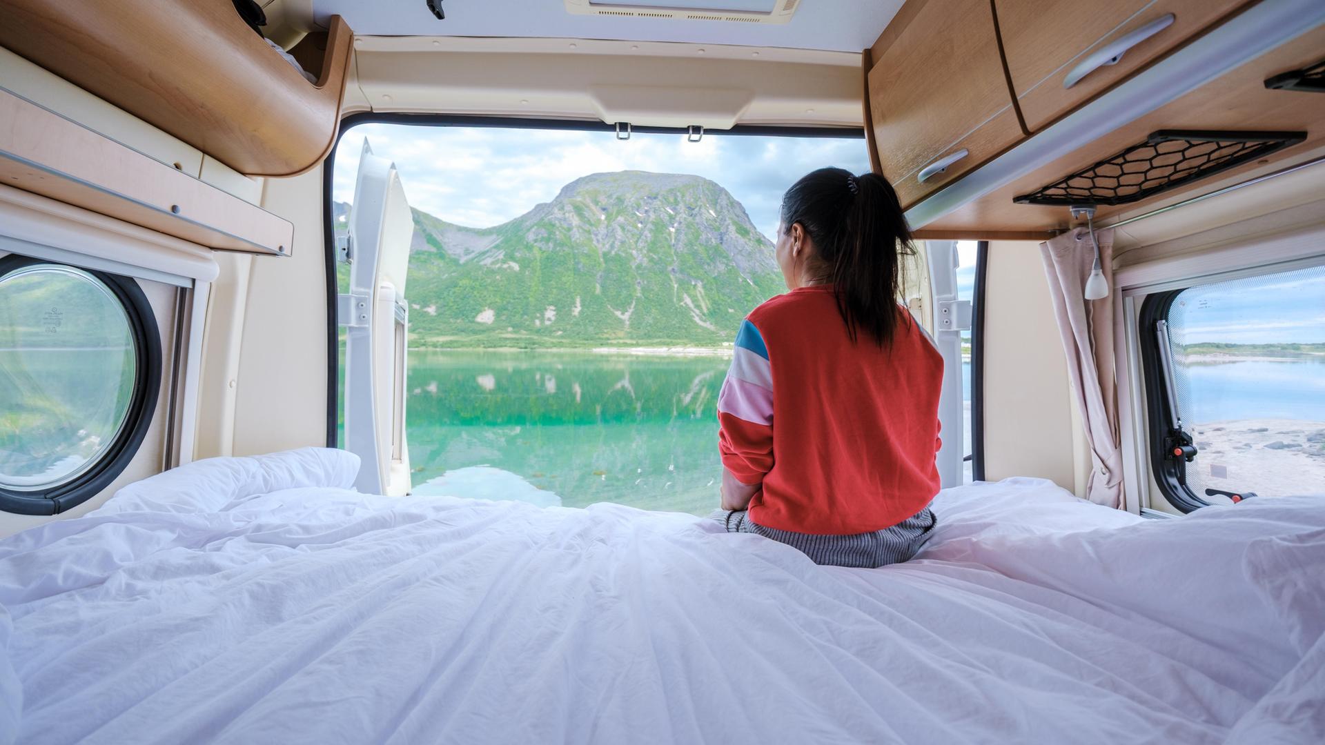 Eine Frau sitzt im Campervan und schaut durch die geöffnete Heckklappe auf ein Fjordpanorama.