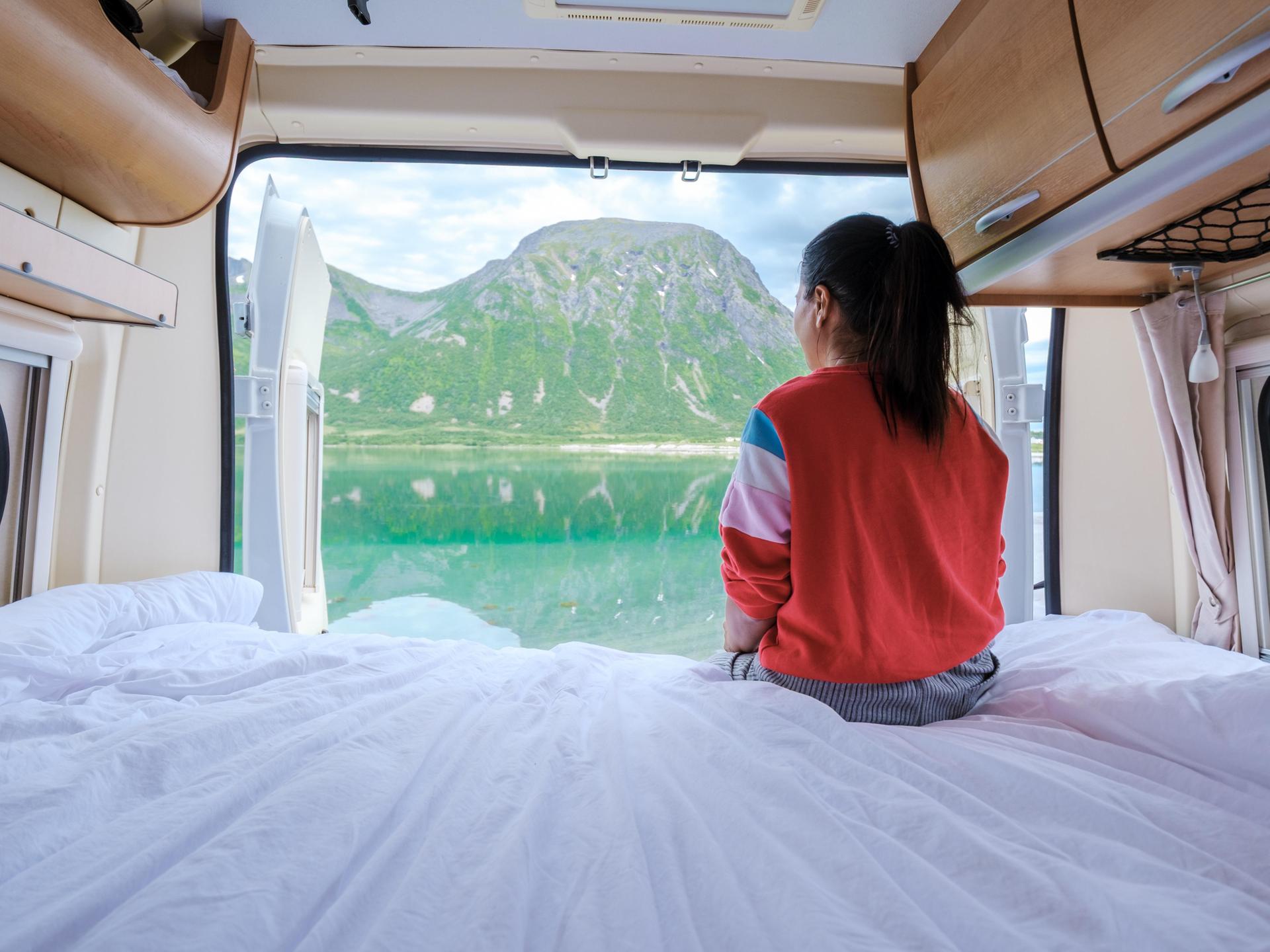 Eine Frau sitzt im Campervan und schaut durch die geöffnete Heckklappe auf ein Fjordpanorama.