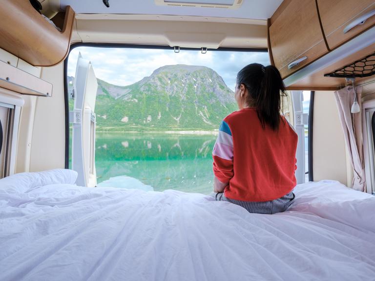 Eine Frau sitzt im Campervan und schaut durch die geöffnete Heckklappe auf ein Fjordpanorama.