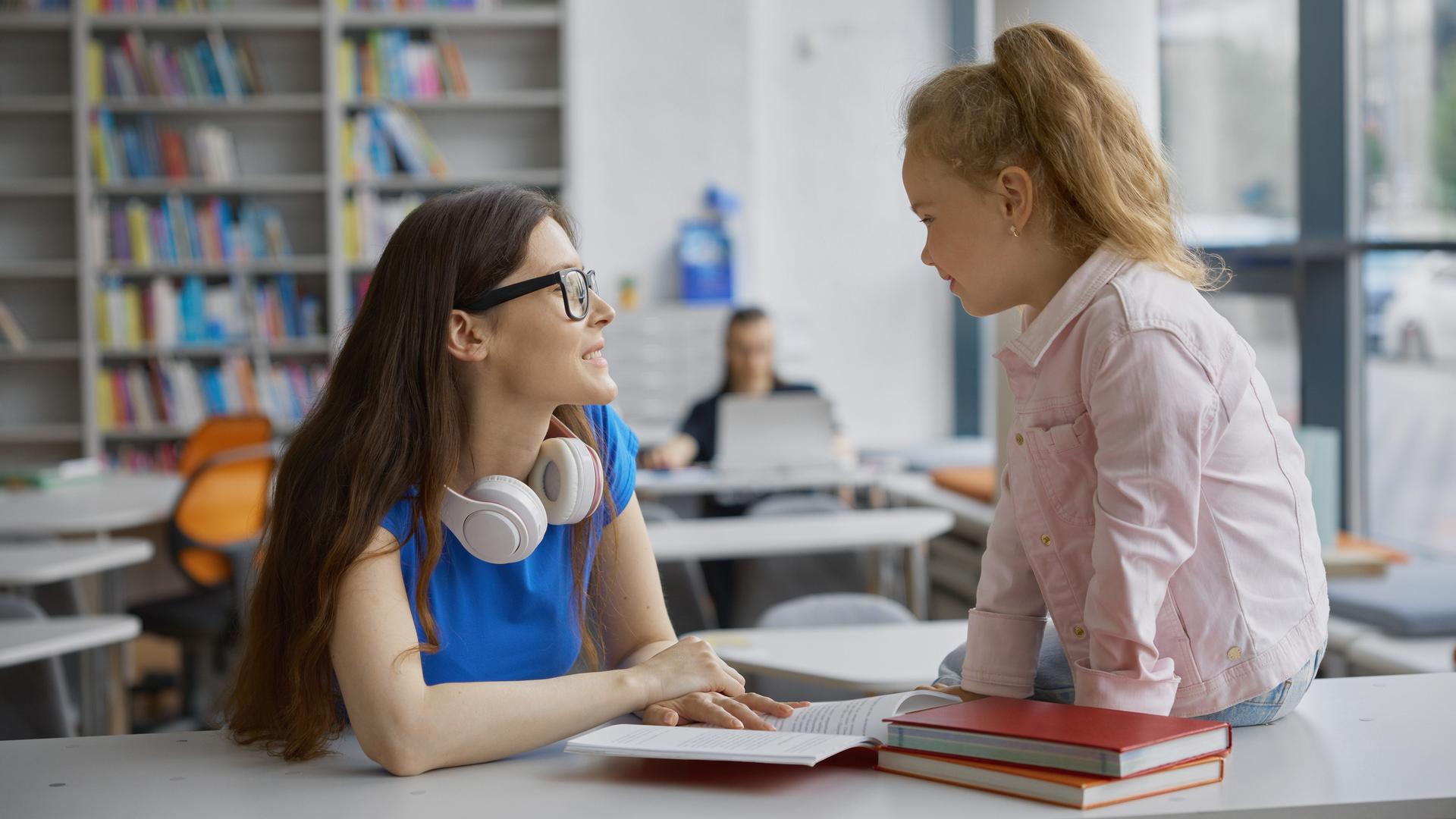 Eine junge Frau spricht in einer Bibliothek mit einem kleinen Mädchen. Das Mädchen sitzt auf dem Tisch. Eine junge Frau spricht in einer Bibliothek mit einem kleinen Mädchen. Das Mädchen sitzt auf dem Tisch.