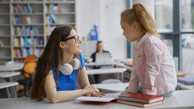 Eine junge Frau spricht in einer Bibliothek mit einem kleinen Mädchen. Das Mädchen sitzt auf dem Tisch.