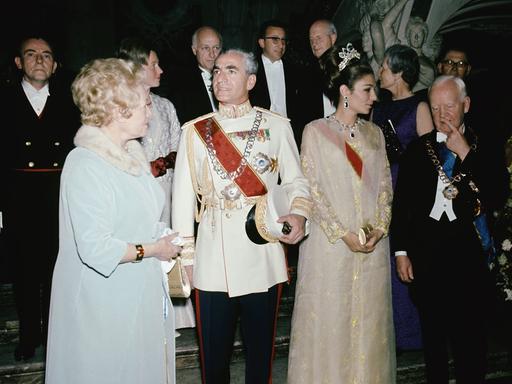 Großer diplomatischer Empfang. Eine ältere Frau im pelzbesetzten Kleid, ein älterer Herr in Uniform, eine jüngere Frau mit einem Krönchen im Haar und ein älterer Herr im Anzug stehen auf einem Teppich.
