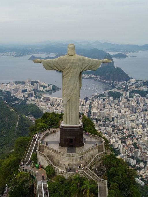 Eine Luftaufnahme zeigt die Stadt Rio De Janeiro umringt von grünen Wäldern und zu Füßen der großen Christus der Erlöser Statue liegend.