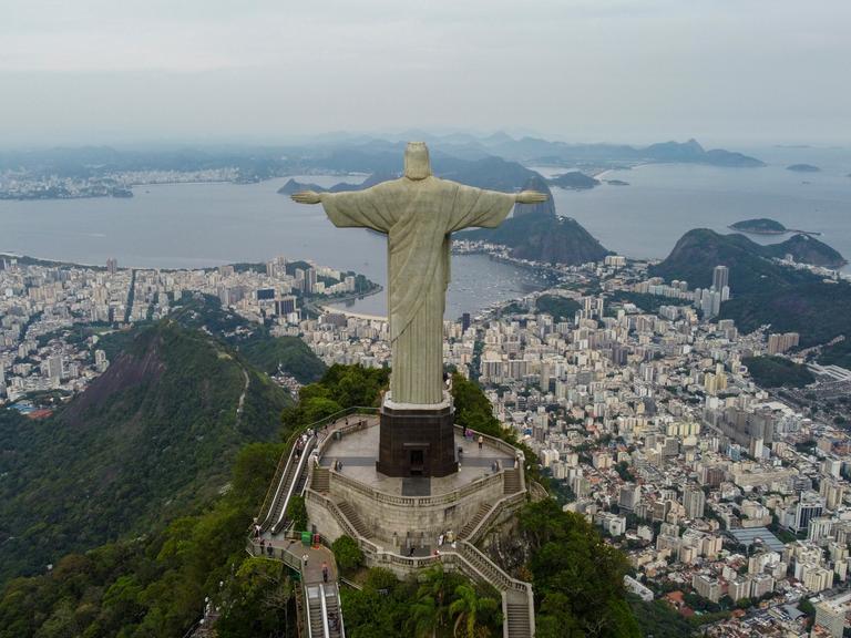 Eine Luftaufnahme zeigt die Stadt Rio De Janeiro umringt von grünen Wäldern und zu Füßen der großen Christus der Erlöser Statue liegend.