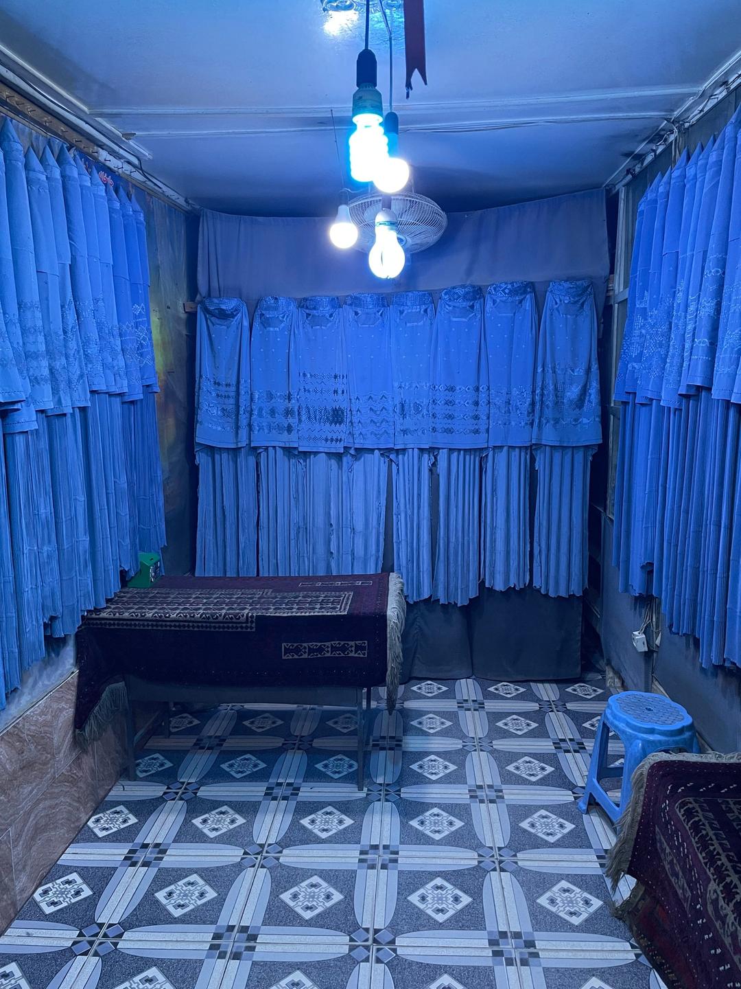 Ein rechteckiger schummrig beleuchteter Raum, der von allen Seiten von dunkelblauen hängenden Burkas eingerahmt wird.