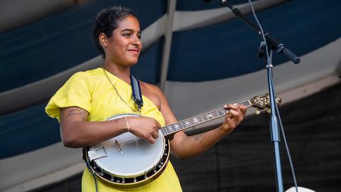 Eine schwarze Musikerin trägt ein gelbes Kleid, steht auf einer Bühne und spielt ein Banjo. 