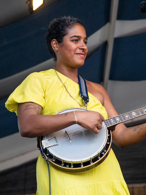 Eine schwarze Musikerin trägt ein gelbes Kleid, steht auf einer Bühne und spielt ein Banjo. 
