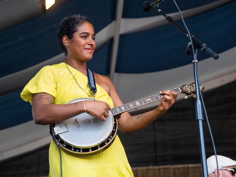 Eine schwarze Musikerin trägt ein gelbes Kleid, steht auf einer Bühne und spielt ein Banjo. 