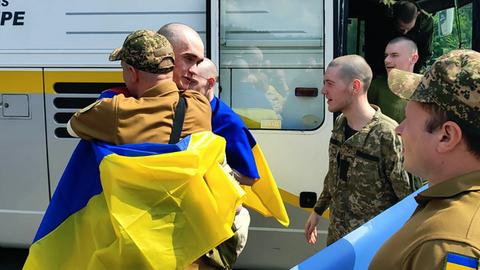 Ein ukrainischer Kriegsgefangener hat eine ukrainische Fahne um die Schultern. Er umarmt einen anderen Mann. Aus einem Bud steigen weitere Männer in Militäruniform.